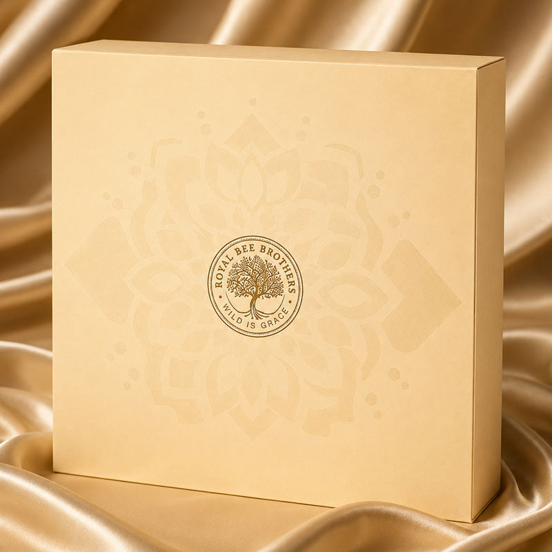 A square cream-colored Royal Bee Brothers gift box with a gold tree of life logo, set against a draped gold satin fabric background.