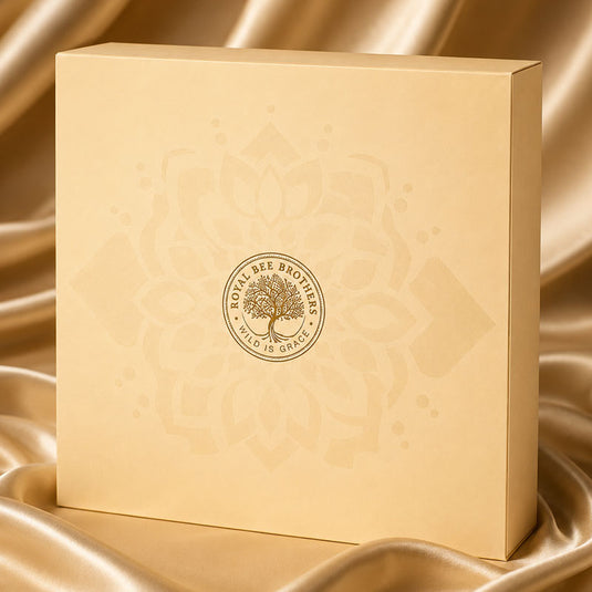 A square cream-colored Royal Bee Brothers gift box with a gold tree of life logo, set against a draped gold satin fabric background.