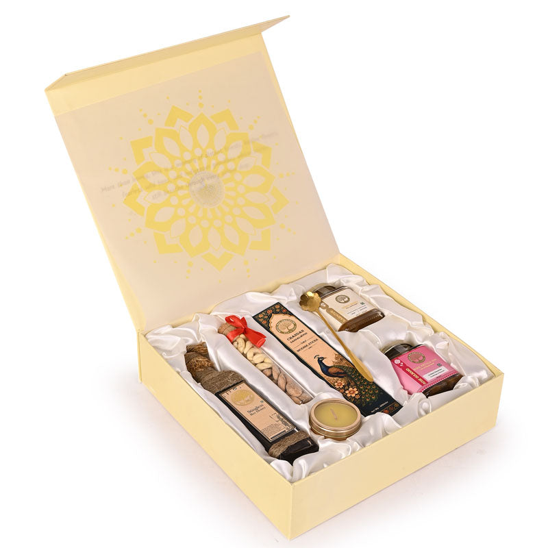 An open cream-colored gift box with a yellow mandala design on the inside lid, containing a curated assortment of premium items including chocolates, a gold-toned candle, a pink packaged item, and a small round tin, all arranged on white satin lining.