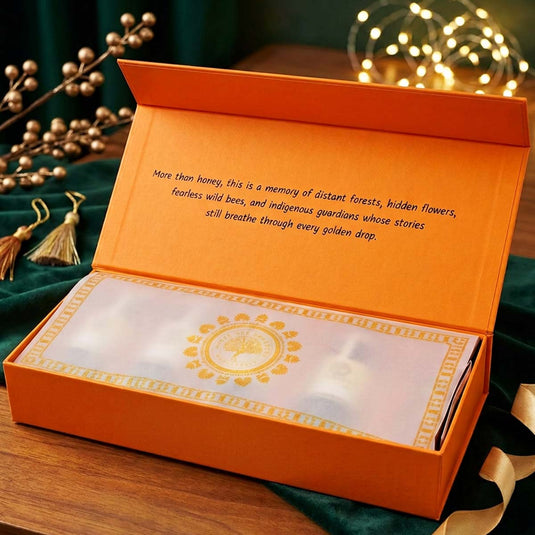 An open orange gift box displayed on a wooden surface, with a translucent white insert showing a decorative gold circular emblem and small white items inside. The inner lid has printed text, and the scene is styled with green fabric, tassel ornaments, and warm string lights in the background.