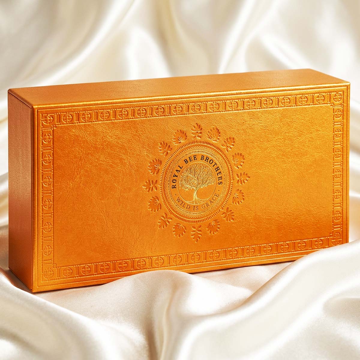 An ornate rectangular golden-orange box with embossed decorative borders and a central Royal Bee Bronzes logo, resting on draped white satin fabric.
