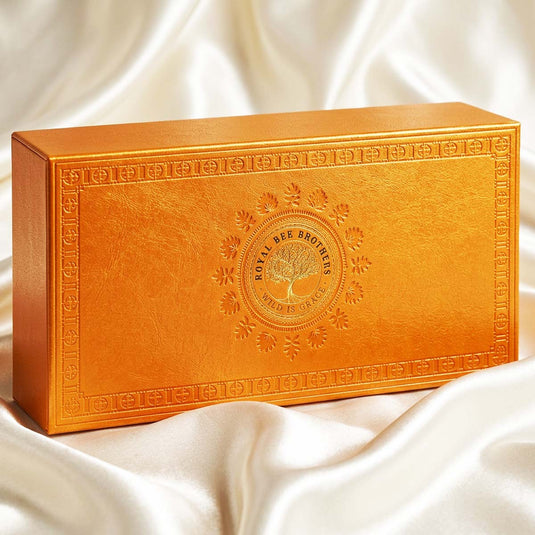 An ornate rectangular golden-orange box with embossed decorative borders and a central Royal Bee Bronzes logo, resting on draped white satin fabric.